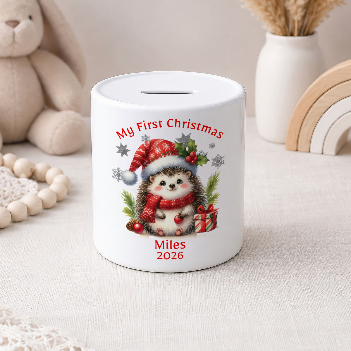 A white ceramic piggy bank with a custom Christmas hedgehog illustration and the text 'My First Christmas' and 'Miles 2026'. The piggy bank is styled on a light beige fabric surface with a plush bunny, wooden beads, a white vase with dried grass, and a wooden rainbow in the background.