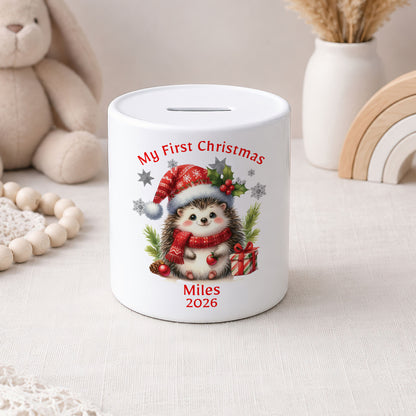 A white ceramic piggy bank with a custom Christmas hedgehog illustration and the text 'My First Christmas' and 'Miles 2026'. The piggy bank is styled on a light beige fabric surface with a plush bunny, wooden beads, a white vase with dried grass, and a wooden rainbow in the background.