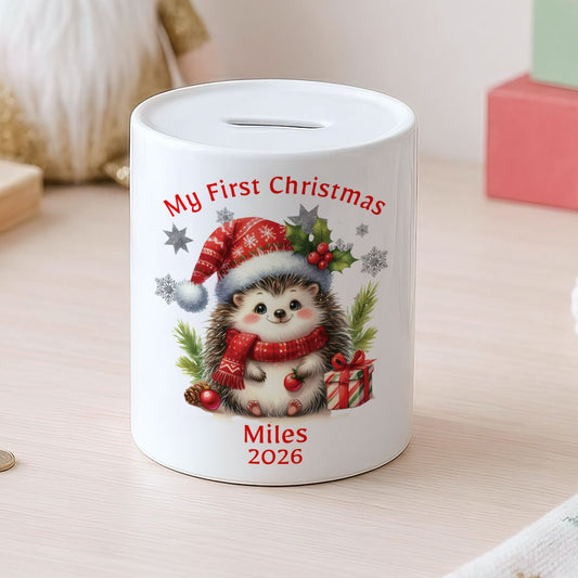 A white ceramic pig savings bank decorated with a cute hedgehog wearing a red Santa hat and scarf, with the text 'My First Christmas', 'Miles 2026', and small silver star accents. The pig bank is set on a light wooden table in a cozy, styled room with a gnome, coins, and colorful blocks in the background.
