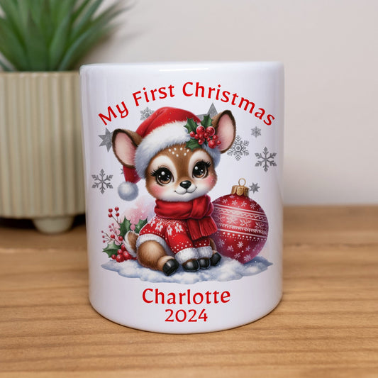 White mug with Christmas-themed design of a baby reindeer, text 'My First Christmas' and 'Charlotte 2024', on a wooden surface with a plant in the background.