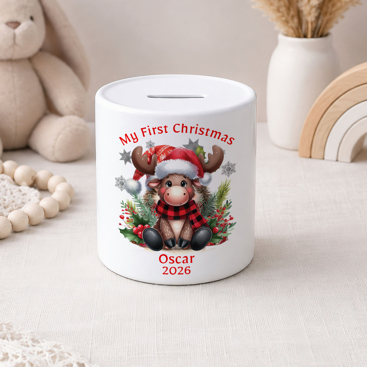 A white ceramic pig savings bank decorated with a festive Christmas design featuring a cartoon reindeer in a red and black plaid scarf, surrounded by holly and a Santa hat. The text 'My First Christmas' is printed above the reindeer, and 'Oscar 2026' is printed below.