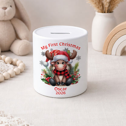 A white ceramic pig savings bank decorated with a festive Christmas design featuring a cartoon reindeer in a red and black plaid scarf, surrounded by holly and a Santa hat. The text 'My First Christmas' is printed above the reindeer, and 'Oscar 2026' is printed below.