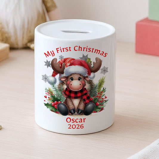 A white ceramic pig savings bank decorated with a festive Christmas illustration of a reindeer wearing a Santa hat and scarf, with the text 'My First Christmas Oscar 2026'. The pig bank is shown on a light wooden table with coins nearby and a cozy, blurred holiday background.