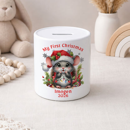 A white ceramic pig bank decorated with a cute cartoon mouse wearing a Santa hat, surrounded by red text reading 'My First Christmas' and 'Imogen 2026'. The bank is styled on a light fabric surface with a plush bunny, wooden beads, and a white vase in the background.