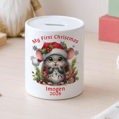 A white ceramic piggy bank with a custom Christmas mouse illustration and the text 'My First Christmas' and 'Imogen 2026', placed on a light wooden table with toys in the background.