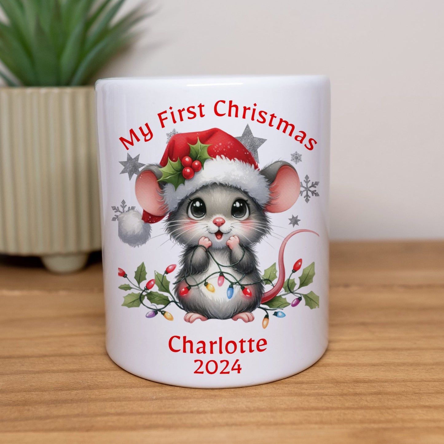 White mug with a Christmas-themed design of a mouse, text 'My First Christmas Charlotte 2024', on a wooden surface with a plant in the background.
