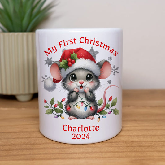 White mug with a Christmas-themed design of a mouse, text 'My First Christmas Charlotte 2024', on a wooden surface with a plant in the background.