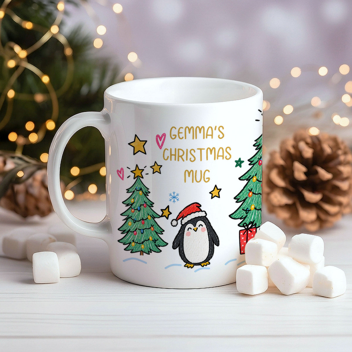 A white ceramic mug with a Christmas design featuring a penguin in a Santa hat, a Christmas tree, stars, and the text 'GEMMA'S CHRISTMAS MUG'. The mug is styled on a white wooden surface with marshmallows, a pinecone, and blurred festive lights in the background.