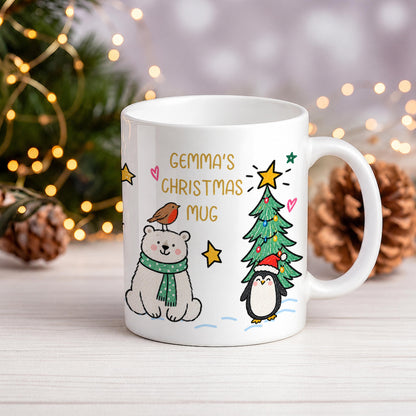 A white ceramic mug with a Christmas-themed illustration featuring a polar bear, penguin, bird, and a Christmas tree, with the text 'GEMMA'S CHRISTMAS MUG'. The mug is set on a light wooden surface with blurred pine branches and warm fairy lights in the background.
