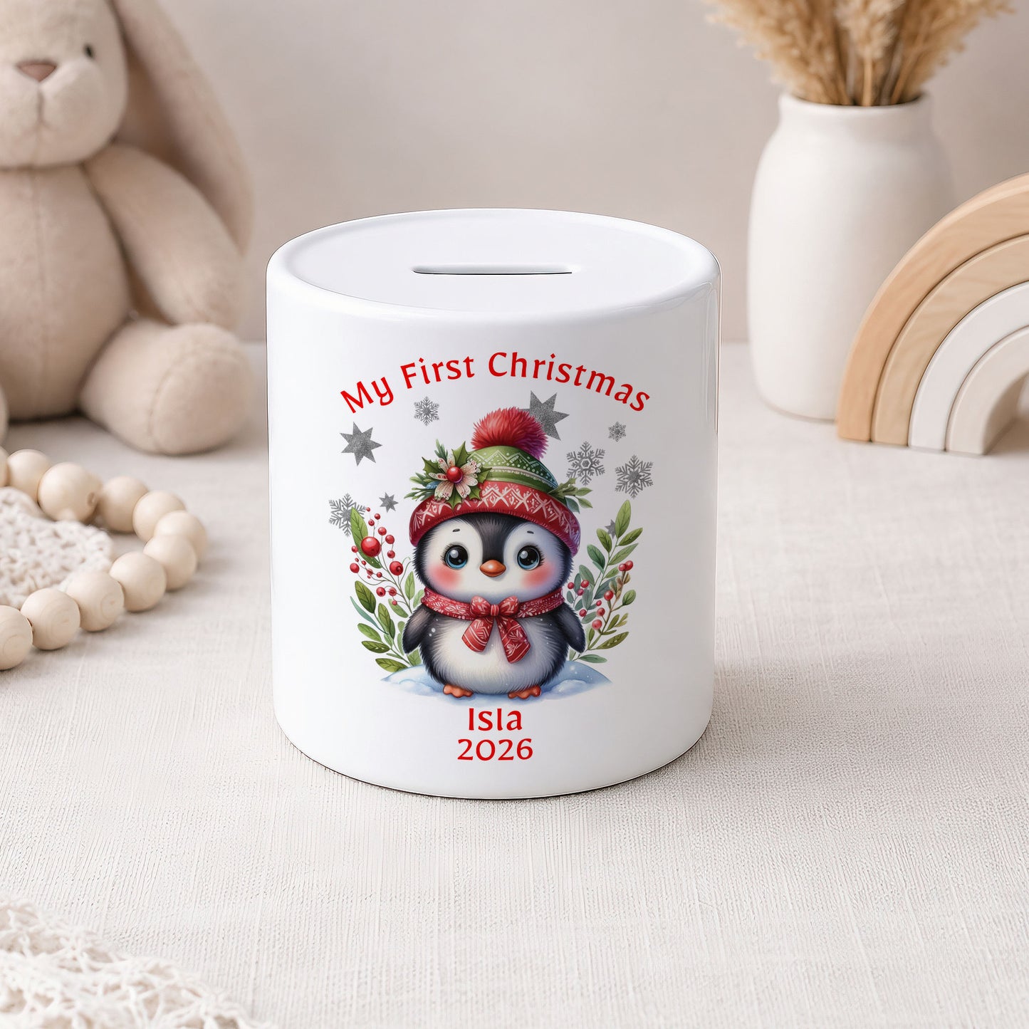 A white ceramic piggy bank with a printed illustration of a penguin wearing a red hat and scarf, surrounded by red berries and green leaves. The text 'My First Christmas' is written above the penguin, and 'Isla 2026' is written below.