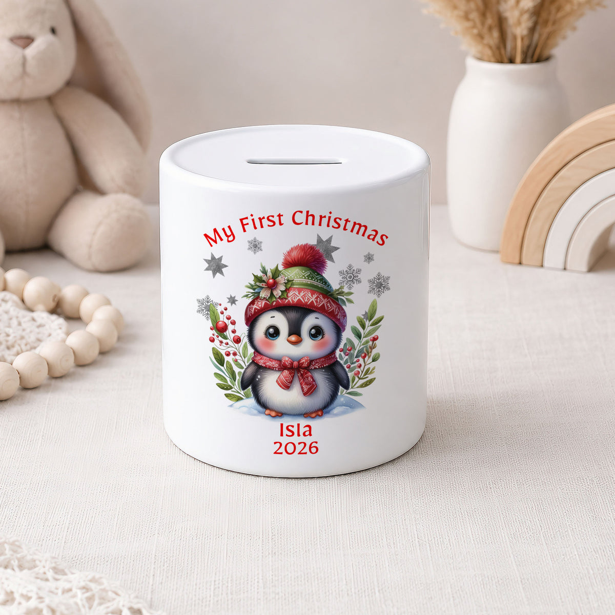 A white ceramic piggy bank with a printed illustration of a penguin wearing a red hat and scarf, surrounded by red berries and green leaves. The text 'My First Christmas' is written above the penguin, and 'Isla 2026' is written below.
