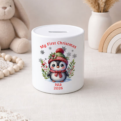 A white ceramic piggy bank with a printed illustration of a penguin wearing a red hat and scarf, surrounded by red berries and green leaves. The text 'My First Christmas' is written above the penguin, and 'Isla 2026' is written below.