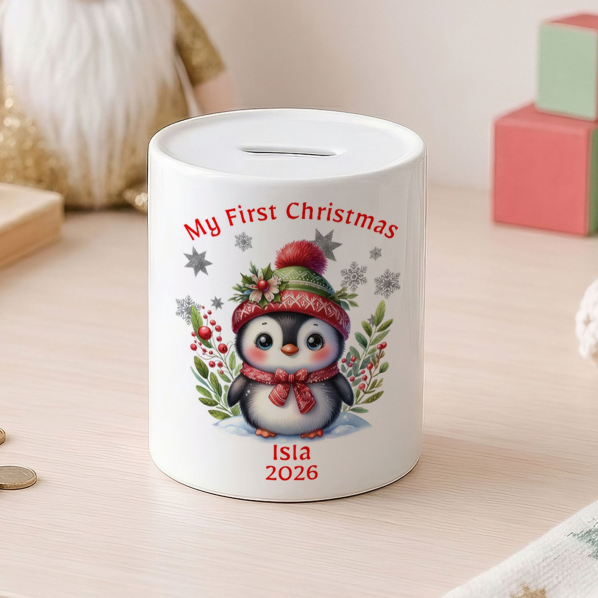A white cylindrical pig bank with a Christmas penguin illustration and the text 'My First Christmas' and 'Isla 2026', set on a light wooden table with coins nearby and soft background decor.