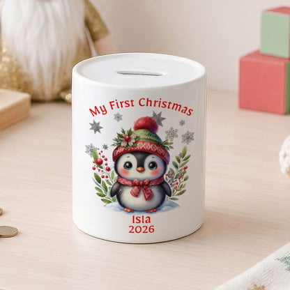 A white cylindrical pig bank with a Christmas penguin illustration and the text 'My First Christmas' and 'Isla 2026', set on a light wooden table with coins nearby and soft background decor.