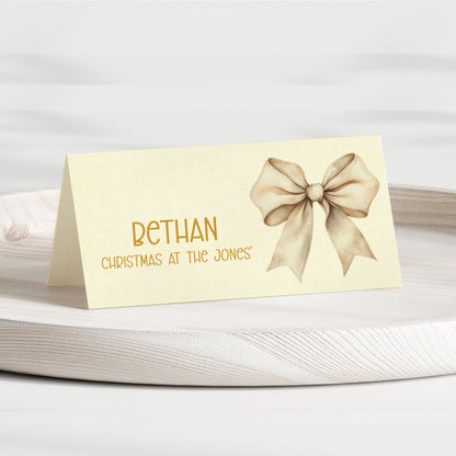 A personalised Christmas table place card with a cream-colored linen texture, featuring the name 'BETHAN' in gold lettering, the phrase 'CHRISTMAS AT THE JONES' below, and a matching gold bow illustration on the right. The card is displayed on a light-colored wooden tray against a soft, neutral background.