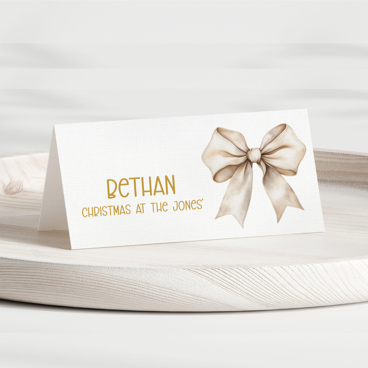 A personalised place card with a white background, featuring the name 'BETHAN' in gold lettering, the phrase 'CHRISTMAS AT THE JONES' below it, and a watercolour-style beige bow on the right side. The card is displayed on a light-coloured wooden tray against a soft, neutral background.