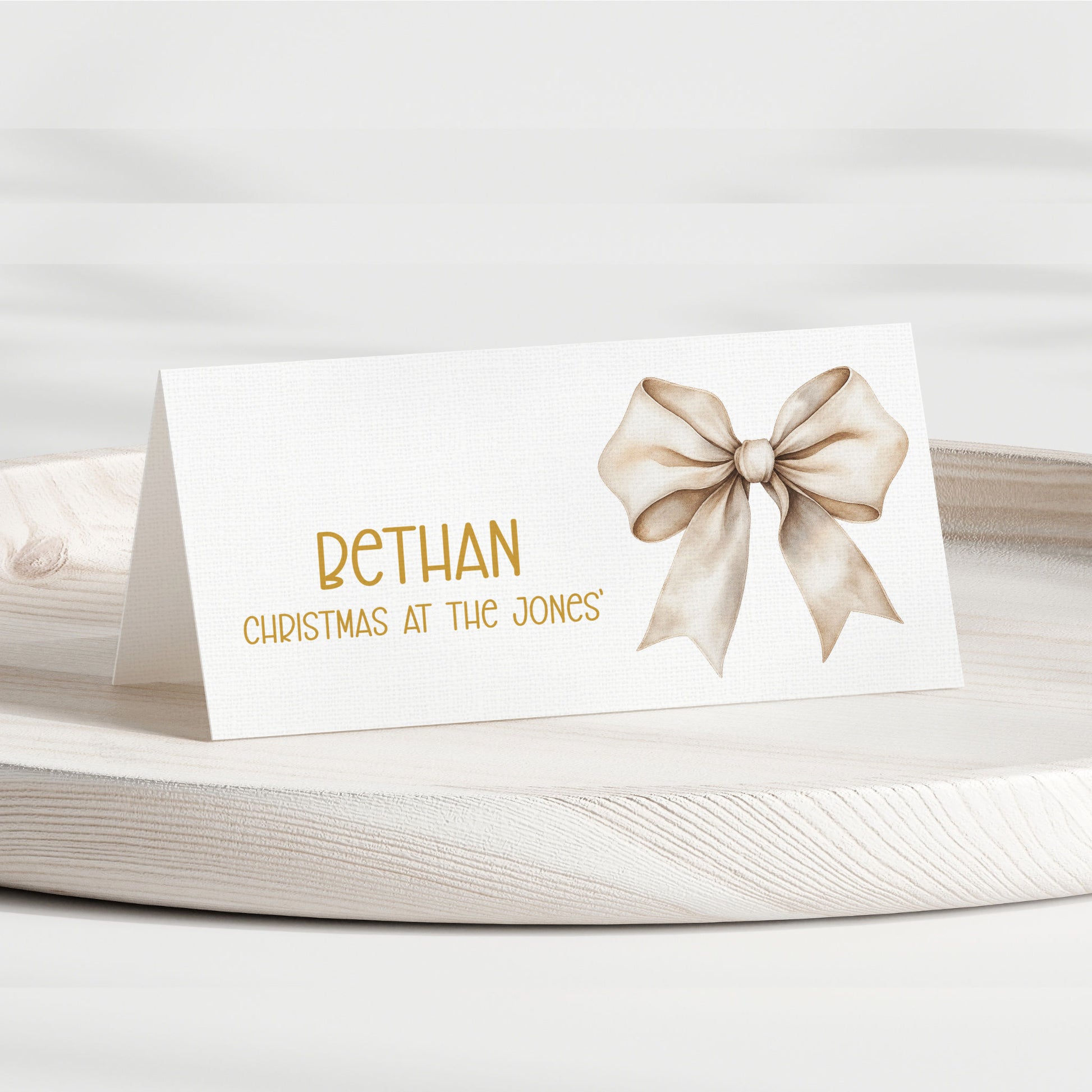 A personalised place card with a white background, featuring the name 'BETHAN' in gold lettering, the phrase 'CHRISTMAS AT THE JONES' below it, and a watercolour-style beige bow on the right side. The card is displayed on a light-coloured wooden tray against a soft, neutral background.