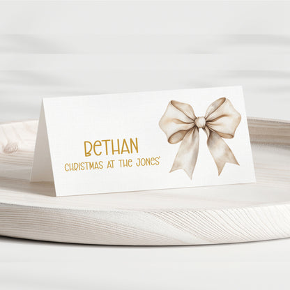 A personalised place card with a white background, featuring the name 'BETHAN' in gold lettering, the phrase 'CHRISTMAS AT THE JONES' below it, and a watercolour-style beige bow on the right side. The card is displayed on a light-coloured wooden tray against a soft, neutral background.
