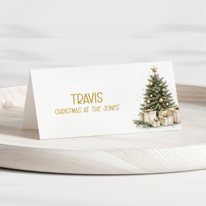 A personalised place card with a white background, featuring the name 'TRAVIS' in gold lettering, the phrase 'CHRISTMAS AT THE JONES' below it, and a watercolour-style Christmas tree on the right side. The card is displayed on a light-coloured wooden tray against a soft, neutral background.