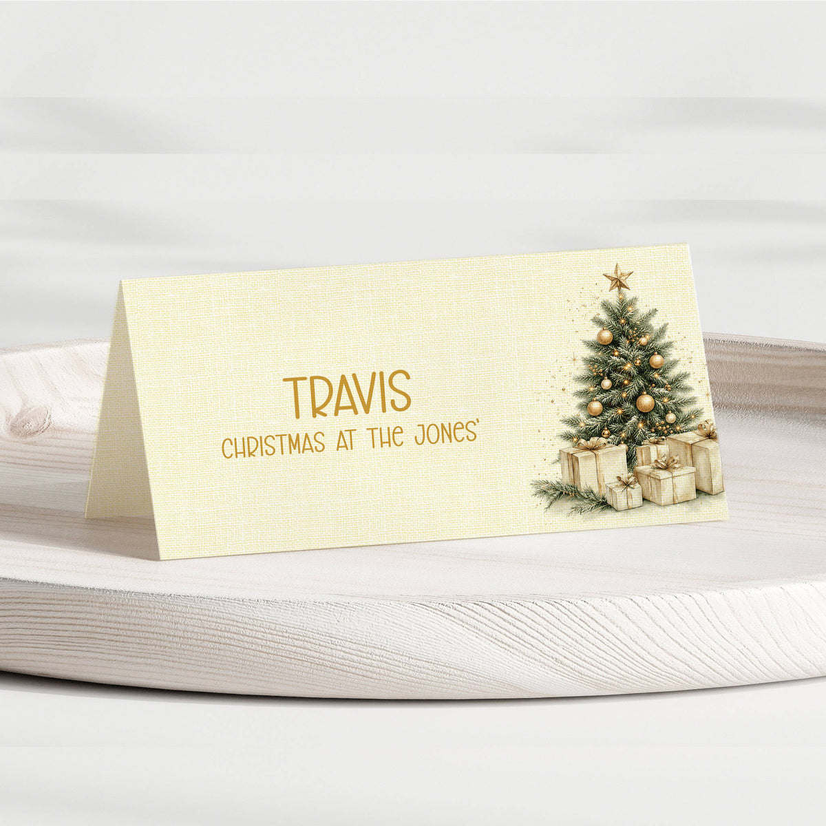 A personalised Christmas table place card with a gold foil name 'TRAVIS' and the phrase 'CHRISTMAS AT THE JONES' on a cream linen-textured background, featuring an illustration of a decorated Christmas tree and wrapped gifts. The card is displayed on a light-colored wooden tray.