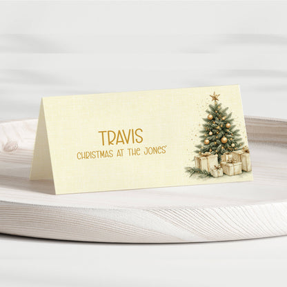 A personalised Christmas table place card with a gold foil name 'TRAVIS' and the phrase 'CHRISTMAS AT THE JONES' on a cream linen-textured background, featuring an illustration of a decorated Christmas tree and wrapped gifts. The card is displayed on a light-colored wooden tray.