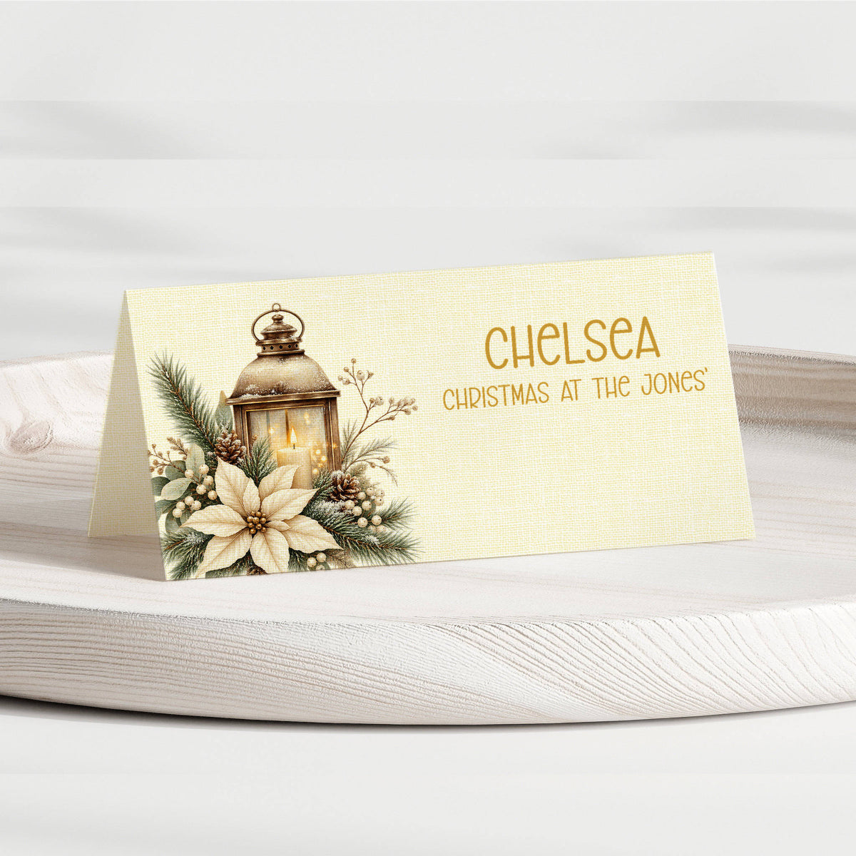 A personalised Christmas place card with a cream linen-textured background, featuring a vintage lantern, pine branches, and white flowers on the left, with the name 'CHELSEA' and the phrase 'CHRISTMAS AT THE JONES' in gold lettering on the right. The card is displayed standing on a white wooden surface.