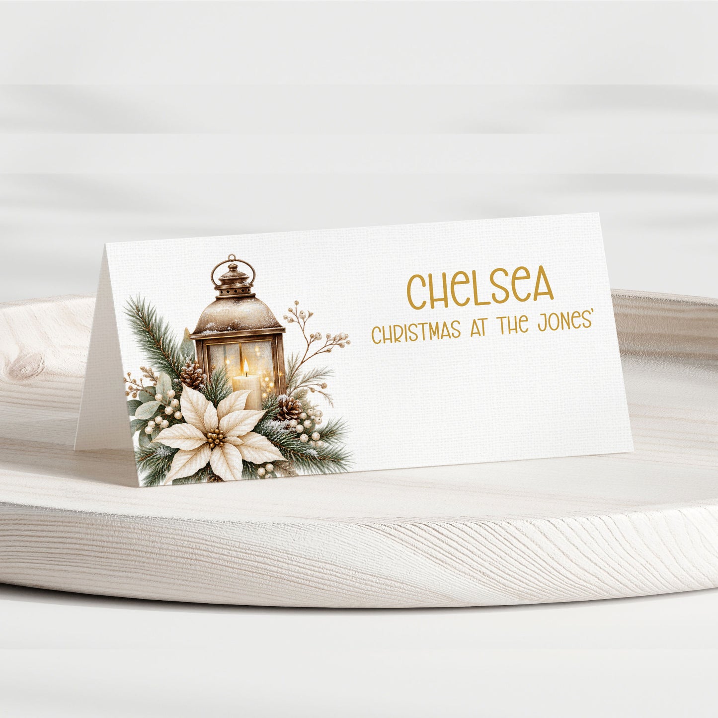 A personalised Christmas table place card on a white wooden tray. The card features a white background with a printed illustration of a lantern, pine branches, berries, and a white flower on the left, and the text 'CHELSEA CHRISTMAS AT THE JONES' in gold on the right.