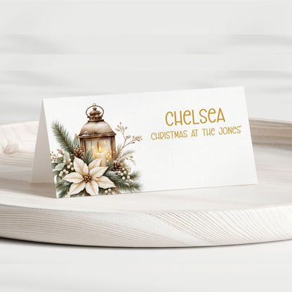 A personalised Christmas table place card on a white wooden tray. The card features a white background with a printed illustration of a lantern, pine branches, berries, and a white flower on the left, and the text 'CHELSEA CHRISTMAS AT THE JONES' in gold on the right.