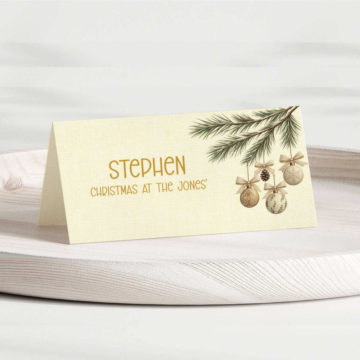 A personalised Christmas place card with a cream linen-textured background, featuring gold text that reads 'STEPHEN CHRISTMAS AT THE JONES' and an illustration of a pine branch with gold ornaments, displayed on a white wooden tray.
