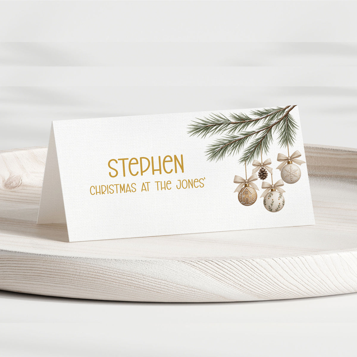 A personalised Christmas table place card with a white background, gold text reading 'STEPHEN CHRISTMAS AT THE JONES', and a pine branch with gold-toned ornaments, displayed on a light wood tray.