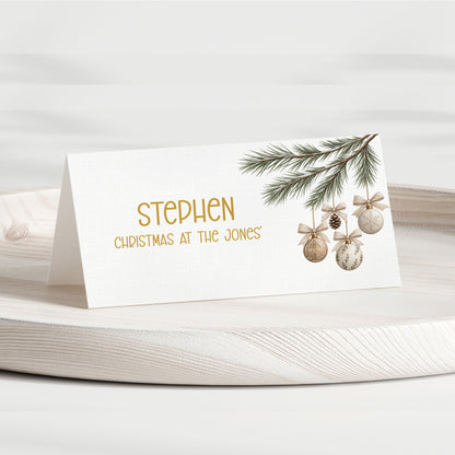 A personalised Christmas table place card with a white background, gold text reading 'STEPHEN CHRISTMAS AT THE JONES', and a pine branch with gold-toned ornaments, displayed on a light wood tray.