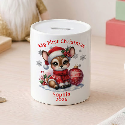A white ceramic pig bank with a Christmas cartoon deer illustration and the text 'My First Christmas Sophie 2026' on the front, shown on a light wooden table with coins nearby and soft-focus holiday decor in the background.