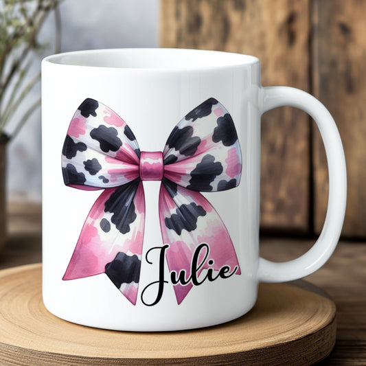 White mug with a pink and black bow design and the name 'Julie' on a wooden coaster.