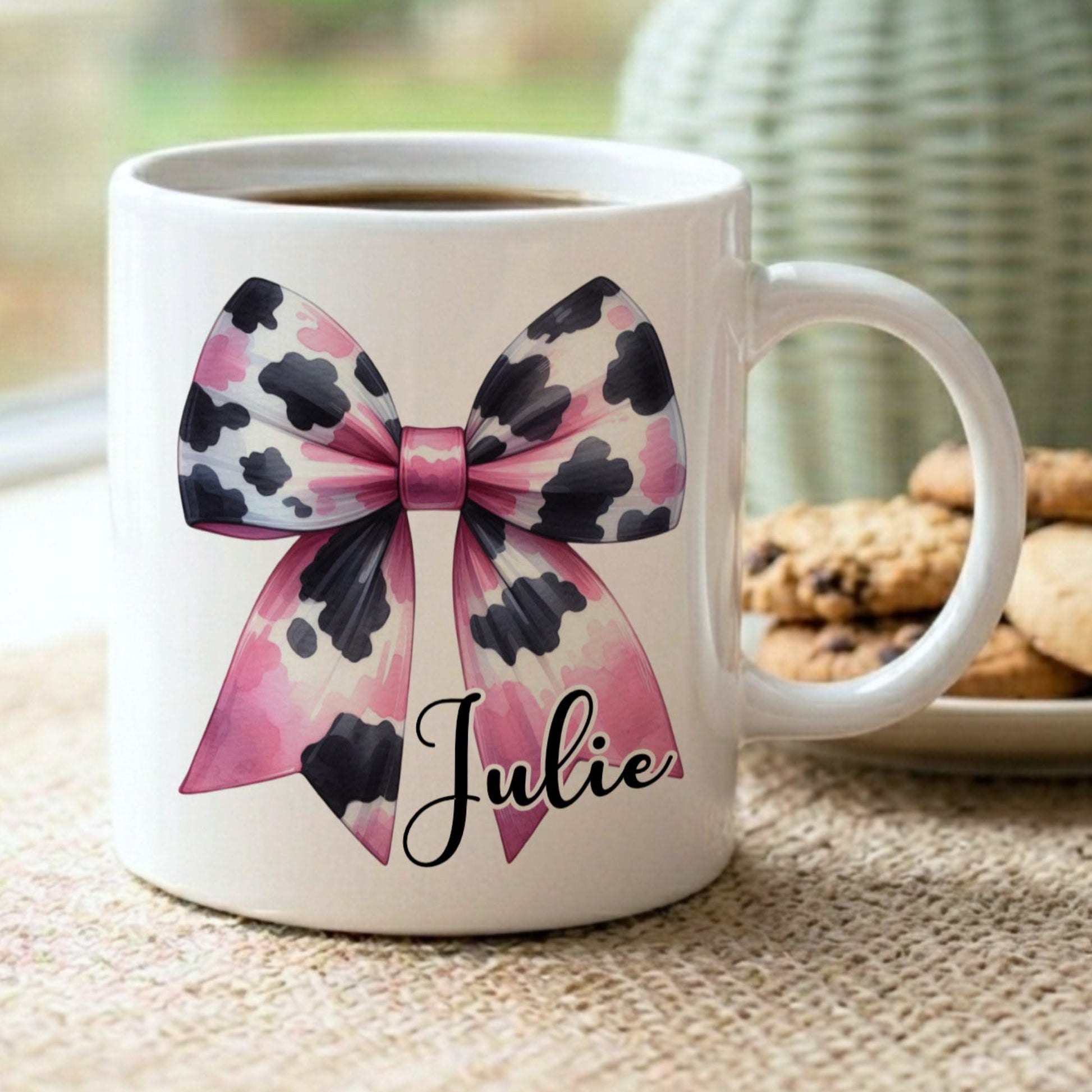 Personalised 'Julie' mug with a pink and black cow print bow design, displayed on a windowsill with cookies and a sage green vase