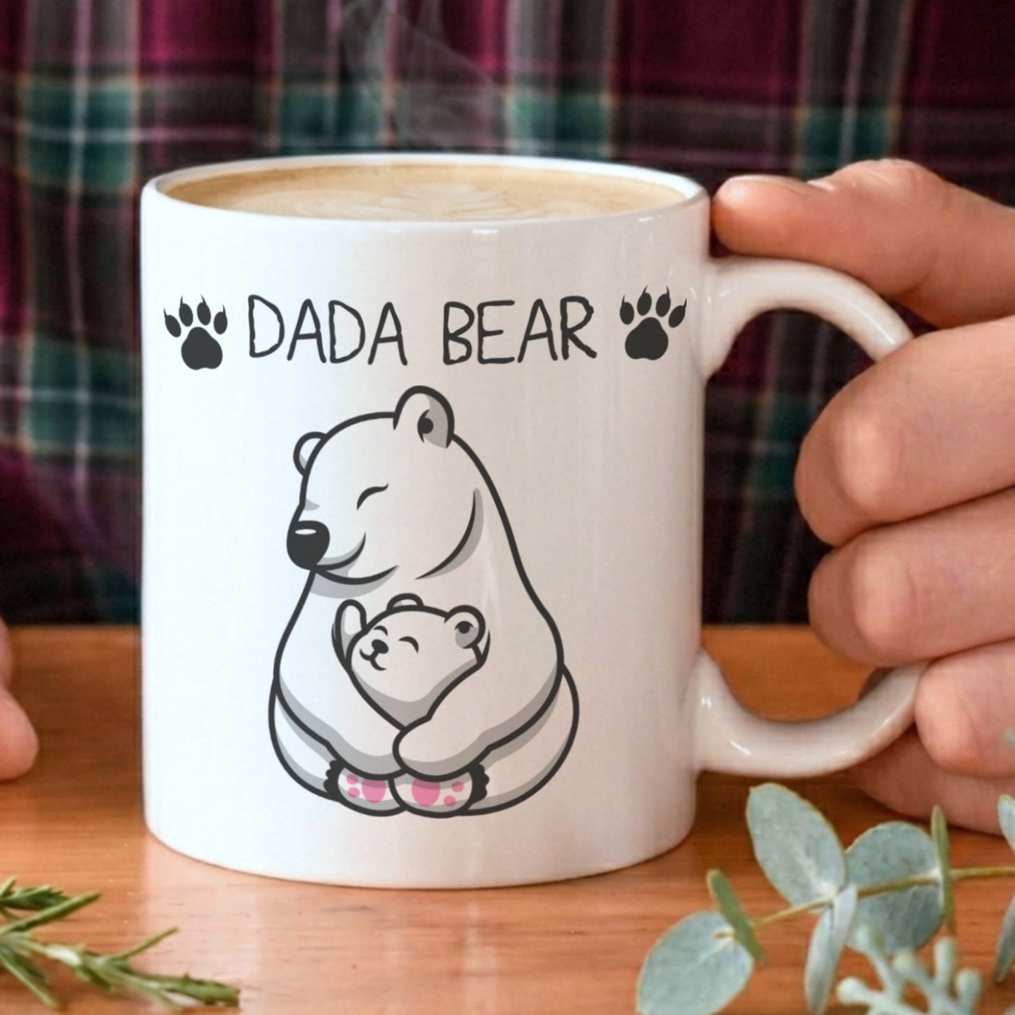 A white ceramic mug with a black line art illustration of a father bear holding a cub and the text 'DADA BEAR' printed on the front. The mug is held by hands against a background of a person wearing a green and red plaid flannel shirt, set on a wooden table with decorative greenery.