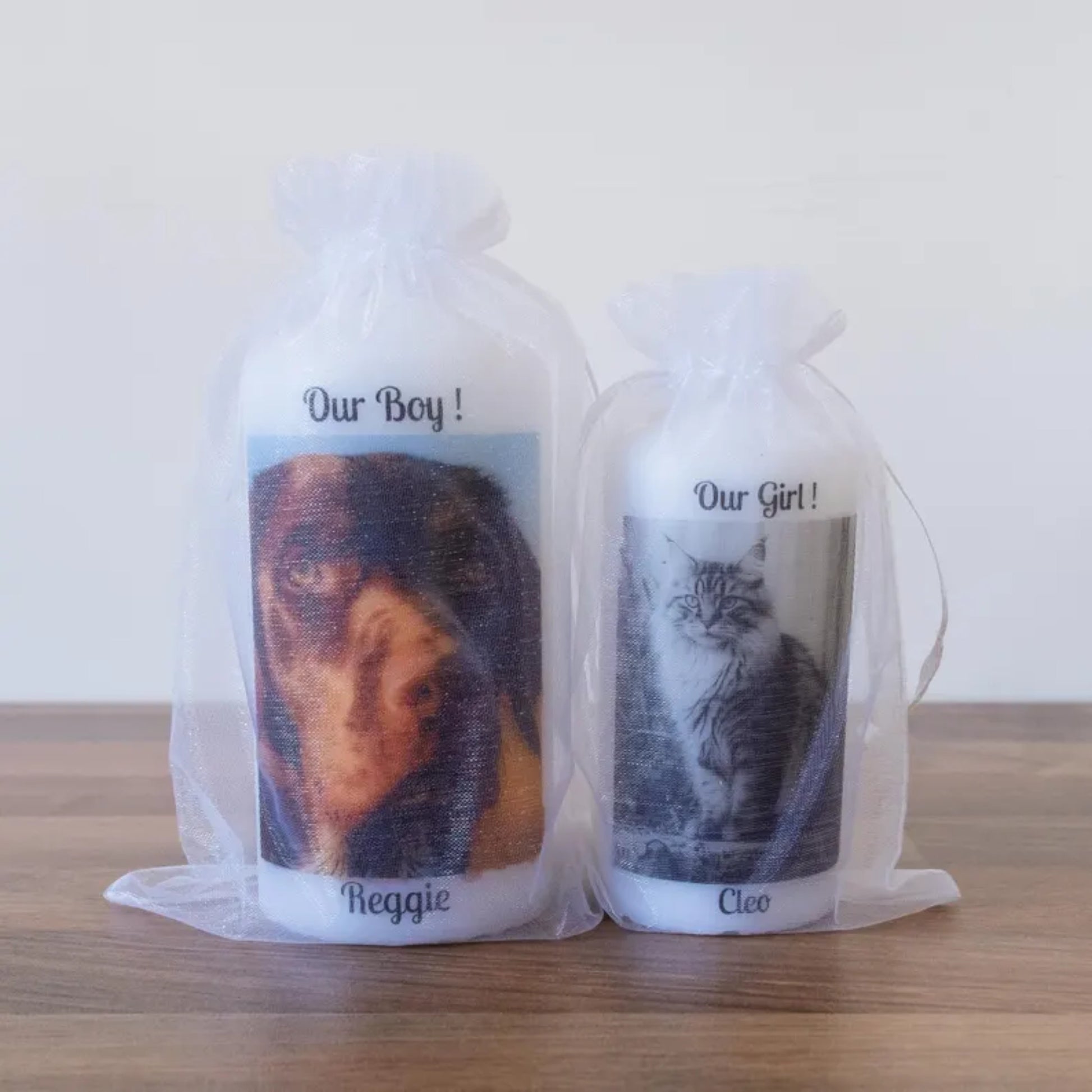 Two white wax candles in clear organza drawstring bags, one printed with a dog portrait labelled 'Our Boy! Reggie' and the other with a cat portrait labelled 'Our Girl! Cleo', displayed on a wooden surface against a plain light background.