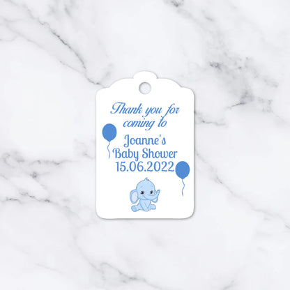 A white baby shower thank you tag with blue text and illustrations of a cartoon elephant and balloons, displayed on a white marble background.