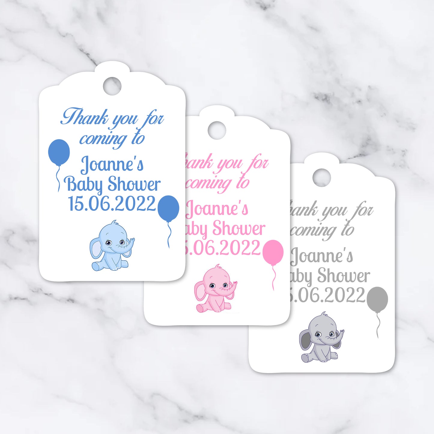 Three white baby shower favour tags with scalloped edges are arranged on a light marble surface. Each tag has a hole at the top and printed text reading 'Thank you for coming to Joanne's Baby Shower 15.