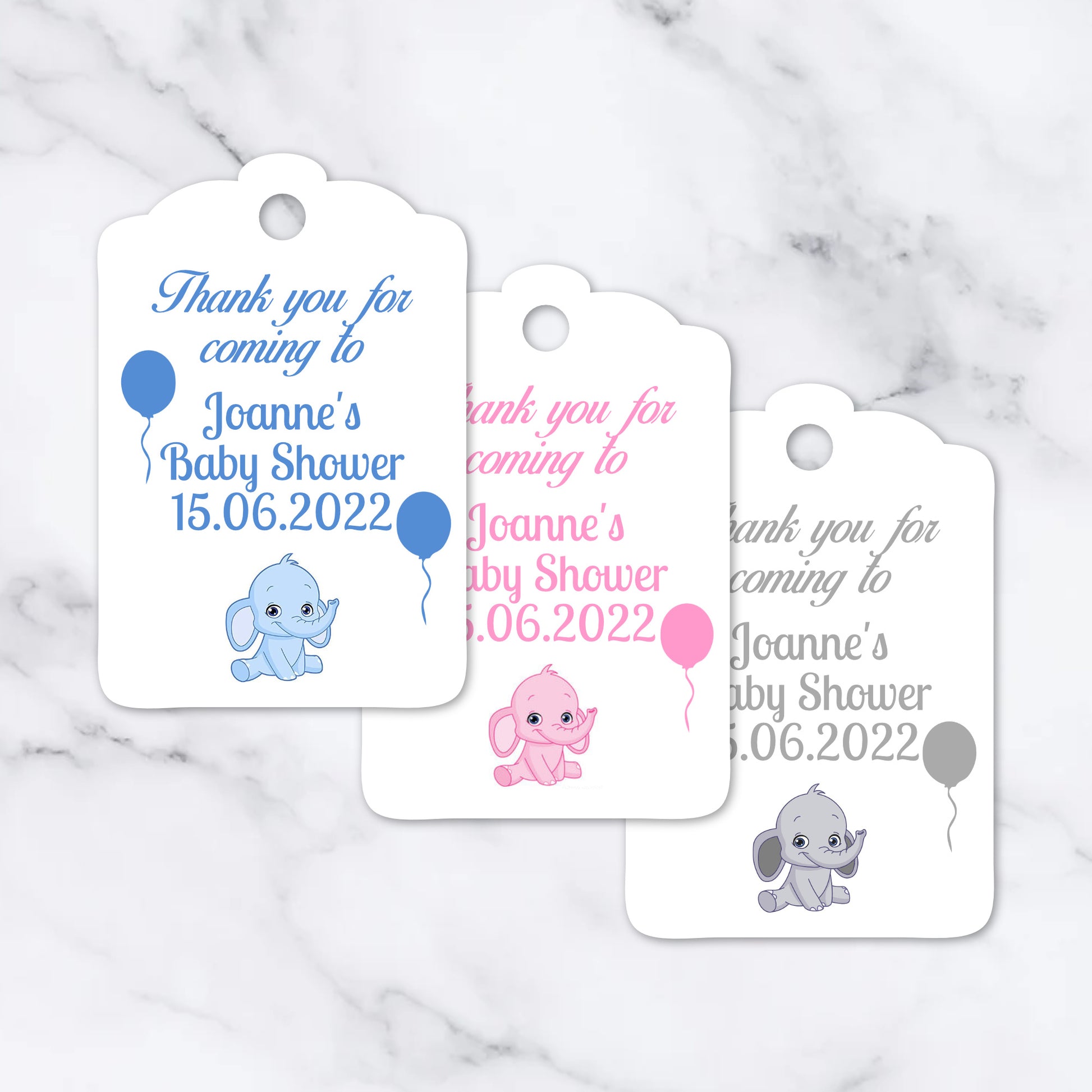 Three white baby shower favour tags with scalloped edges are arranged on a light marble surface. Each tag has a hole at the top and printed text reading 'Thank you for coming to Joanne's Baby Shower 15.