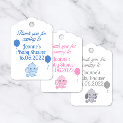 Three white baby shower favour tags with scalloped edges are arranged on a light marble surface. Each tag has a hole at the top and printed text reading 'Thank you for coming to Joanne's Baby Shower 15.