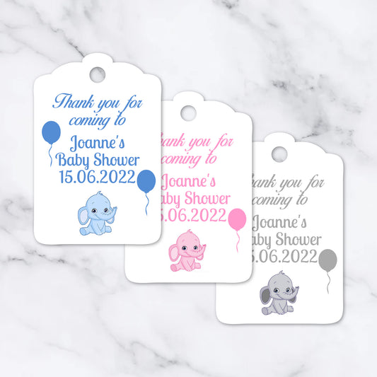 Three white baby shower favour tags with scalloped edges are arranged on a light marble surface. Each tag has a hole at the top and printed text reading 'Thank you for coming to Joanne's Baby Shower 15.
