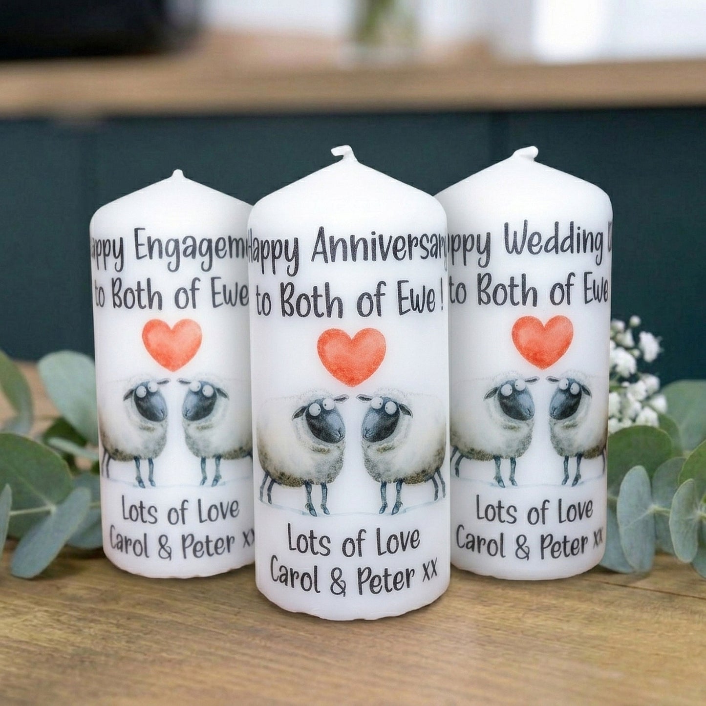 Three white candles standing on a wooden surface, printed with wedding-themed text, an orange heart, and two sheep illustrations. The candles are arranged in front of a dark green background with blurred greenery.