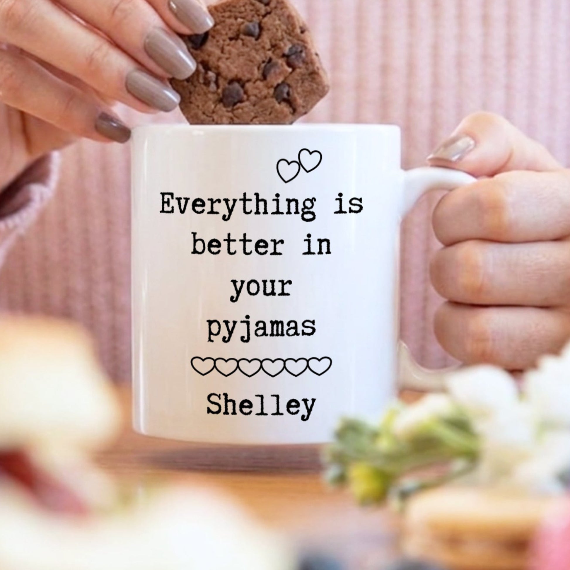 A person in a pink ribbed sweater holds a white mug with the printed phrase 'Everything is better in your pyjamas Shelley' while placing a chocolate chip cookie into the mug. The scene is softly lit and styled with breakfast items in the foreground.