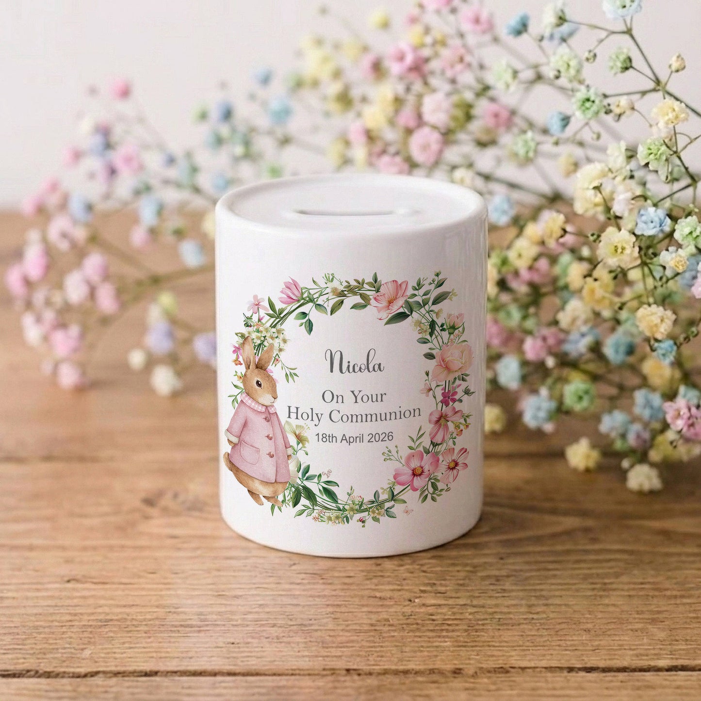 A white ceramic money box with a personalised floral design featuring a bunny and the text 'Nicola On Your Holy Communion 18th April 2026', set on a wooden table with baby's breath flowers in the background.