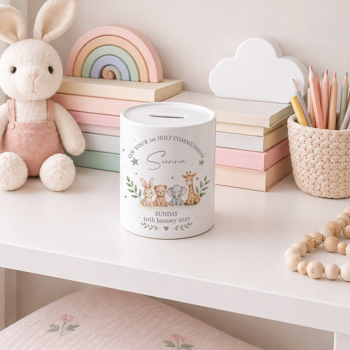 A white cylindrical pig bank with a slot on top, decorated with a cute illustration of a bunny, mouse, and giraffe, and the text 'In Your Holy Communion Sarah Sunday 16th January 2021'. The pig bank is styled on a white shelf next to a plush bunny, pastel books, a wooden rainbow, and a pencil cup.
