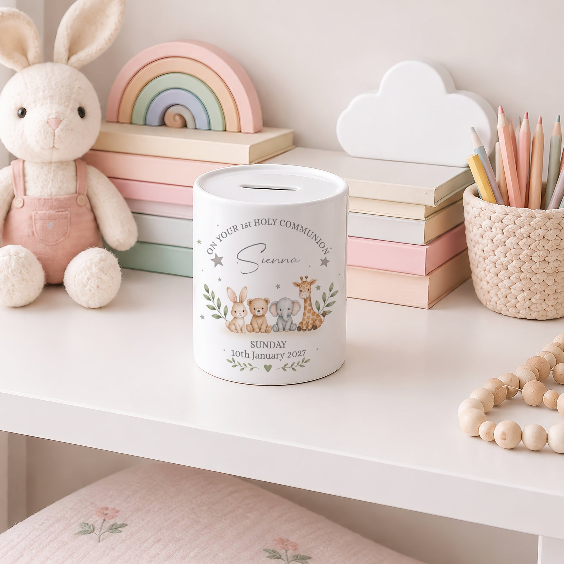 A white cylindrical pig bank with a slot on top, decorated with a cute illustration of a bunny, mouse, and giraffe, and the text 'In Your Holy Communion Sarah Sunday 16th January 2021'. The pig bank is styled on a white shelf next to a plush bunny, pastel books, a wooden rainbow, and a pencil cup.