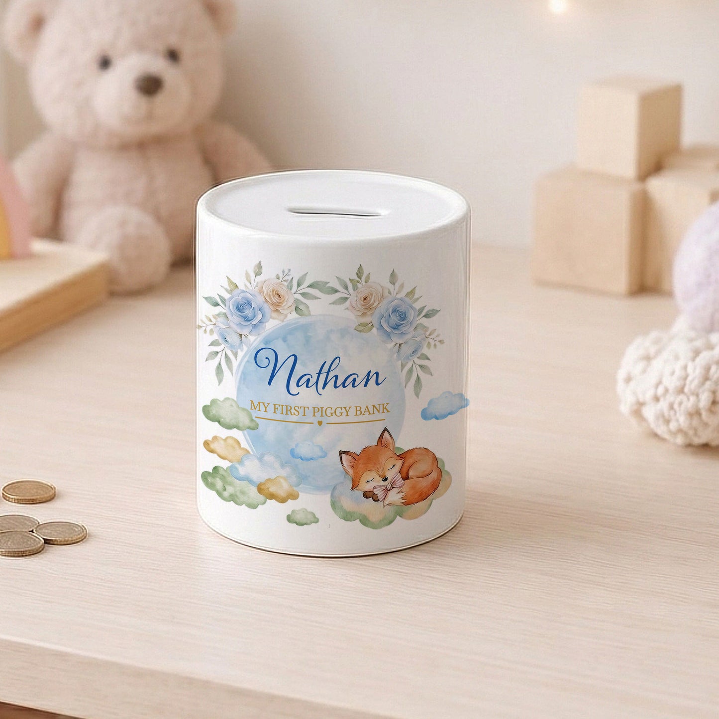 A white cylindrical piggy bank with a floral and fox illustration, personalised with the name 'Nathan' and the text 'MY FIRST PIGGY BANK', sitting on a light wood table with a teddy bear in the background.