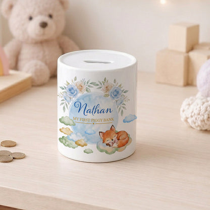 A white cylindrical piggy bank with a floral and fox illustration, personalised with the name 'Nathan' and the text 'MY FIRST PIGGY BANK', sitting on a light wood table with a teddy bear in the background.