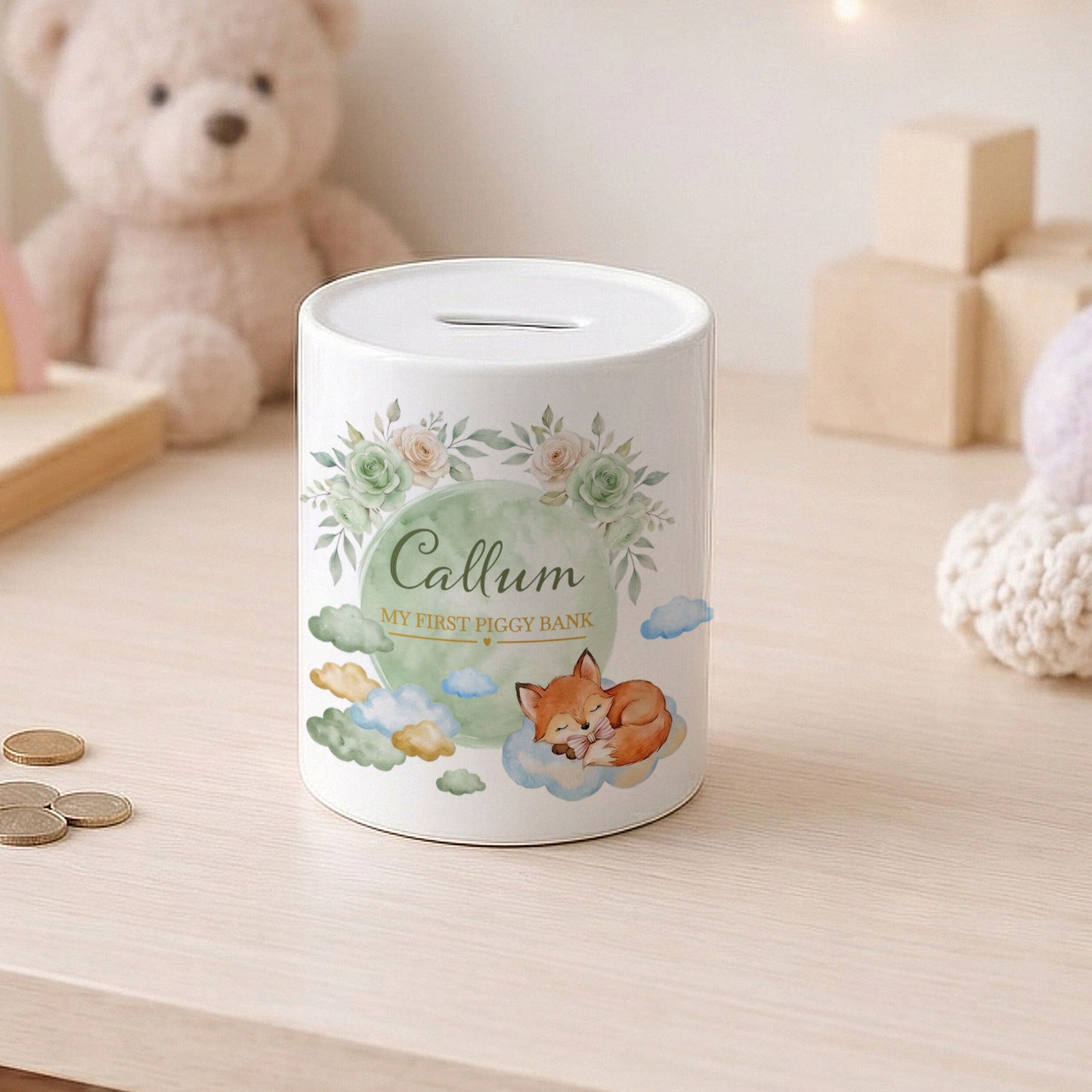 A white ceramic piggy bank with a printed illustration of a sleeping fox, green floral accents, and the name 'Callum' on the front, placed on a light wooden table with a teddy bear in the background.