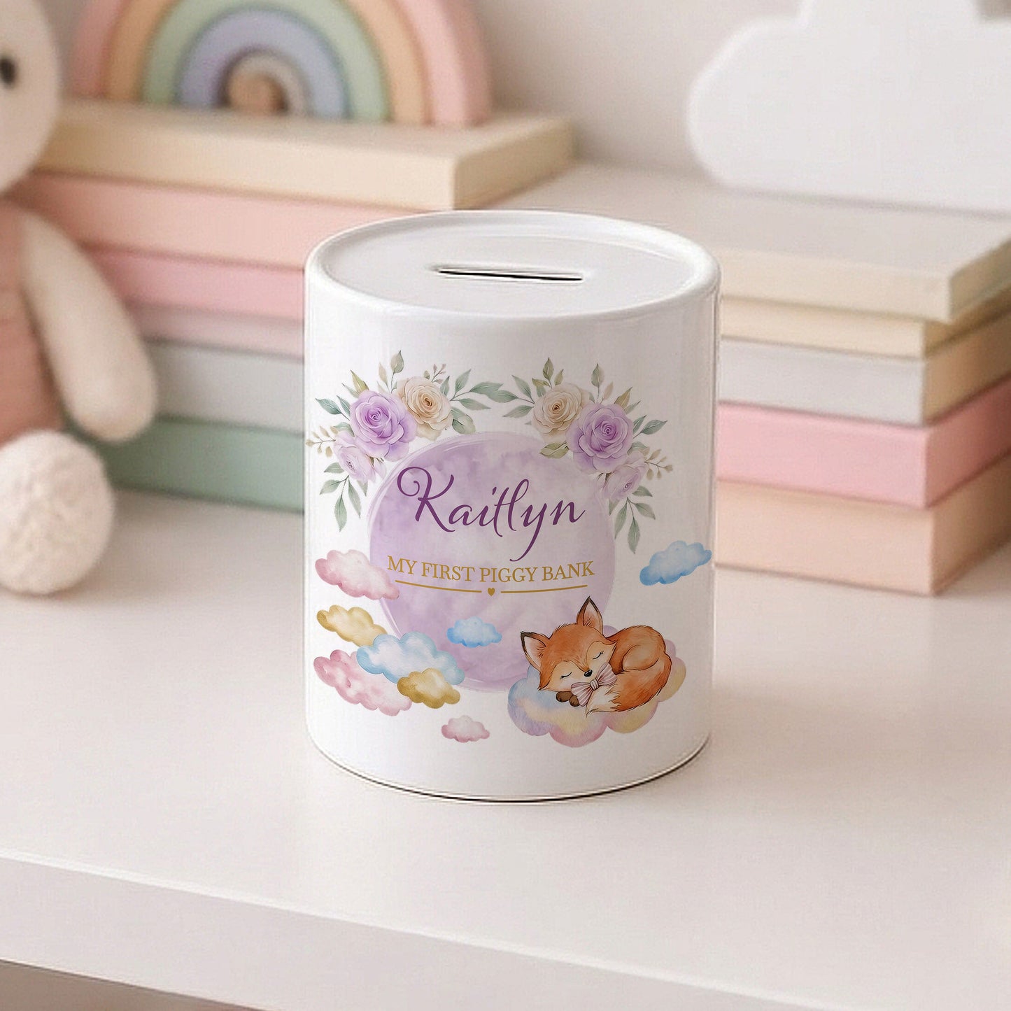 A white cylindrical piggy bank with a slot on top, decorated with pastel floral artwork, a sleeping fox, clouds, and the personalised name 'Kaitlyn' on the front. It sits on a white surface with soft-focus pastel toys and stacked books in the background.